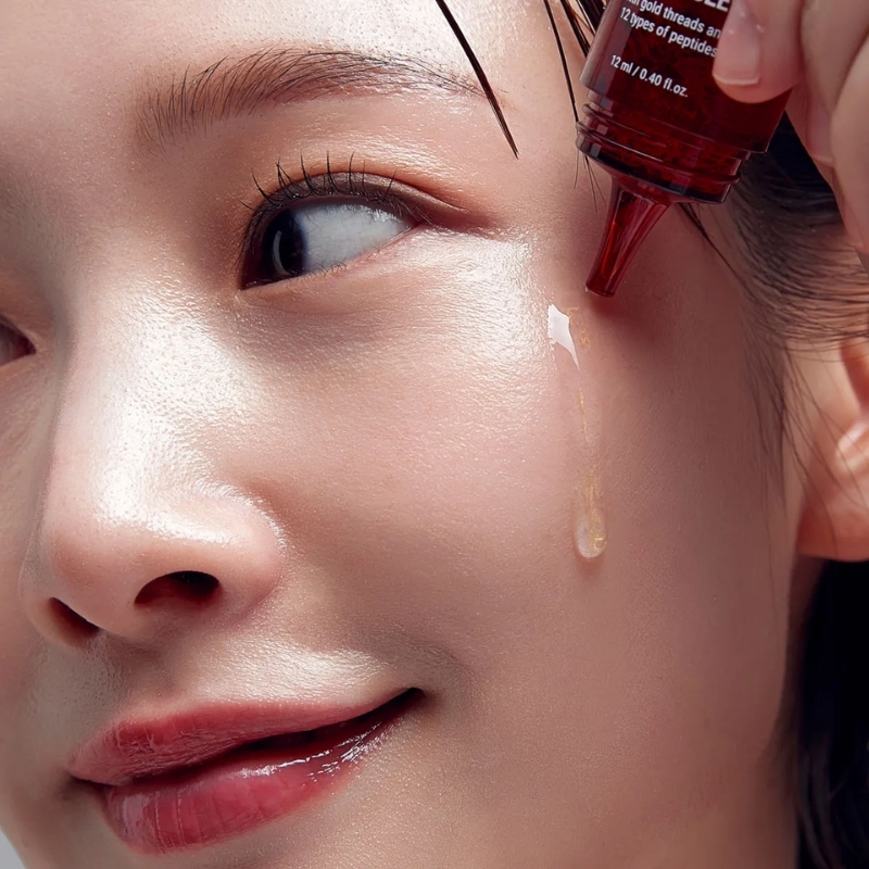 Close-up of a woman's face as she applies amber serum to her cheek with a red dropper.