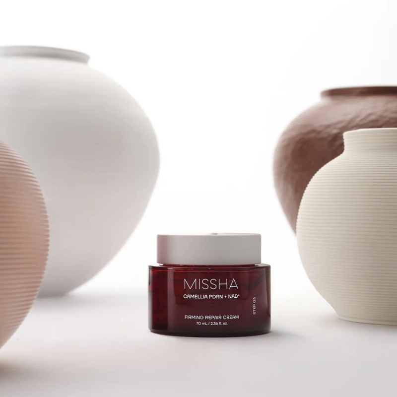 Red MISSHA Firming Repair Cream jar on a white surface, surrounded by cream and white ceramic vases, highlighting the product.